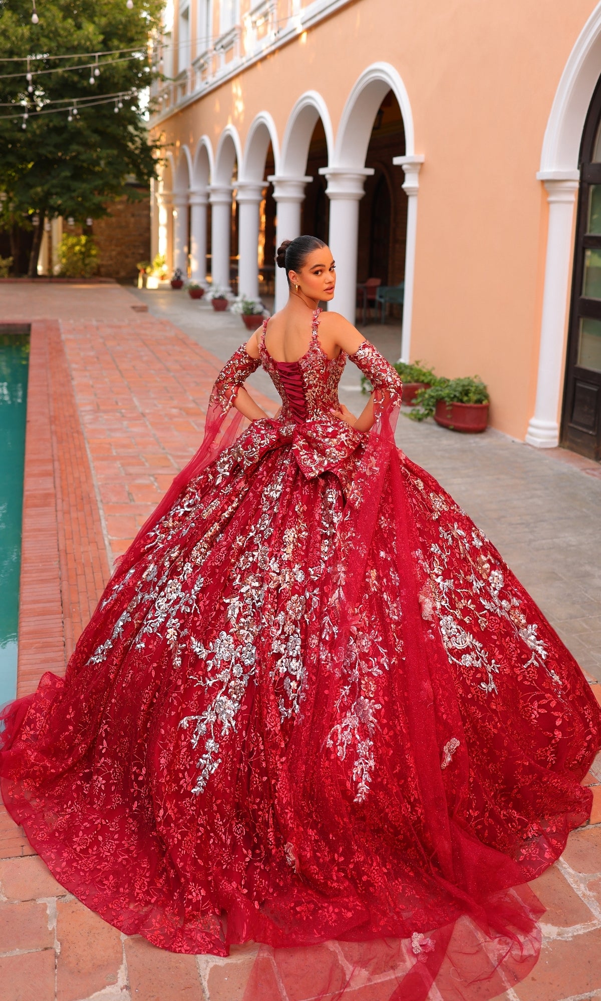 A woman in the Quinceanera Dress 54322 By Amarra stands on a brick pathway beside a pool, with an arched building in the background.